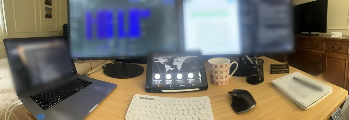 Panorama of a desk: a laptop, two monitors, one showing a calendar packed with blocks, a tablet showing world time zones, a mug, a notebook and pen.