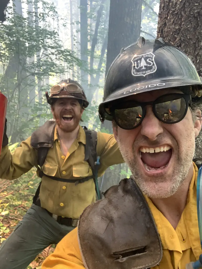 Two of us in yellow shirts and hard hats, grinning in smoky timber. My helmet says LUCIDO.