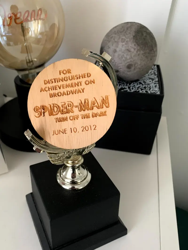 A small trophy on a shelf, its wooden disc engraved: For distinguished achievement on Broadway. Spider-Man, Turn Off the Dark. June 10, 2012.